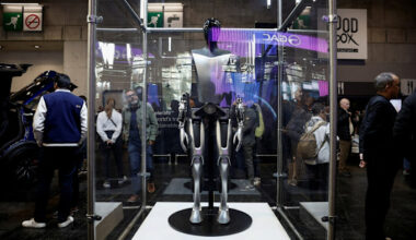 The Tesla Optimus robot is displayed inside a glass case at the 2024 Paris Auto Show, with people observing around it.