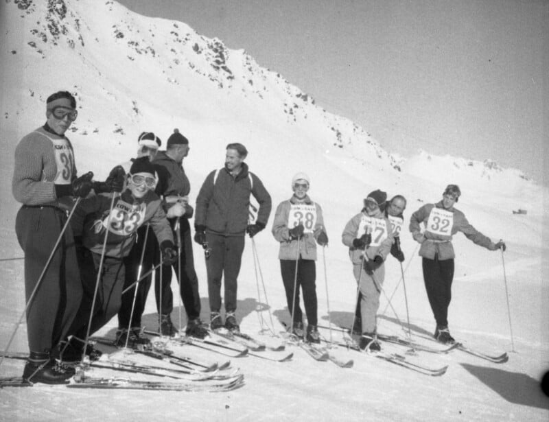 A group of skiers wearing numbered bibs and winter gear stand in a row on a snowy mountain slope, smiling and posing with skis and poles. Snow-covered peaks rise in the background.