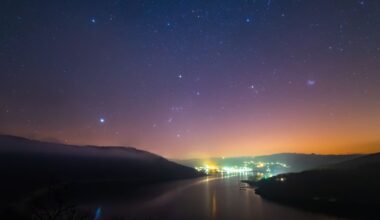 A purple and orange night sky with a few twinkling white stars overlooks distant city lights with a river and two dark banks in the foreground