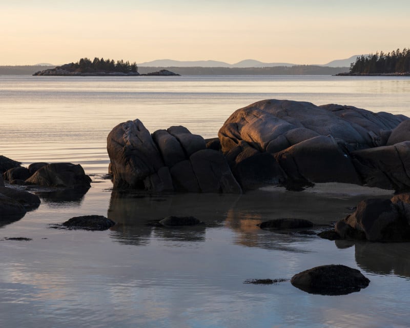 Rocky shoreline at sunset with large boulders in the foreground, calm water, and a tree-covered island in the distance. Soft light casts warm tones on the rocks; mountains are faintly visible on the horizon.