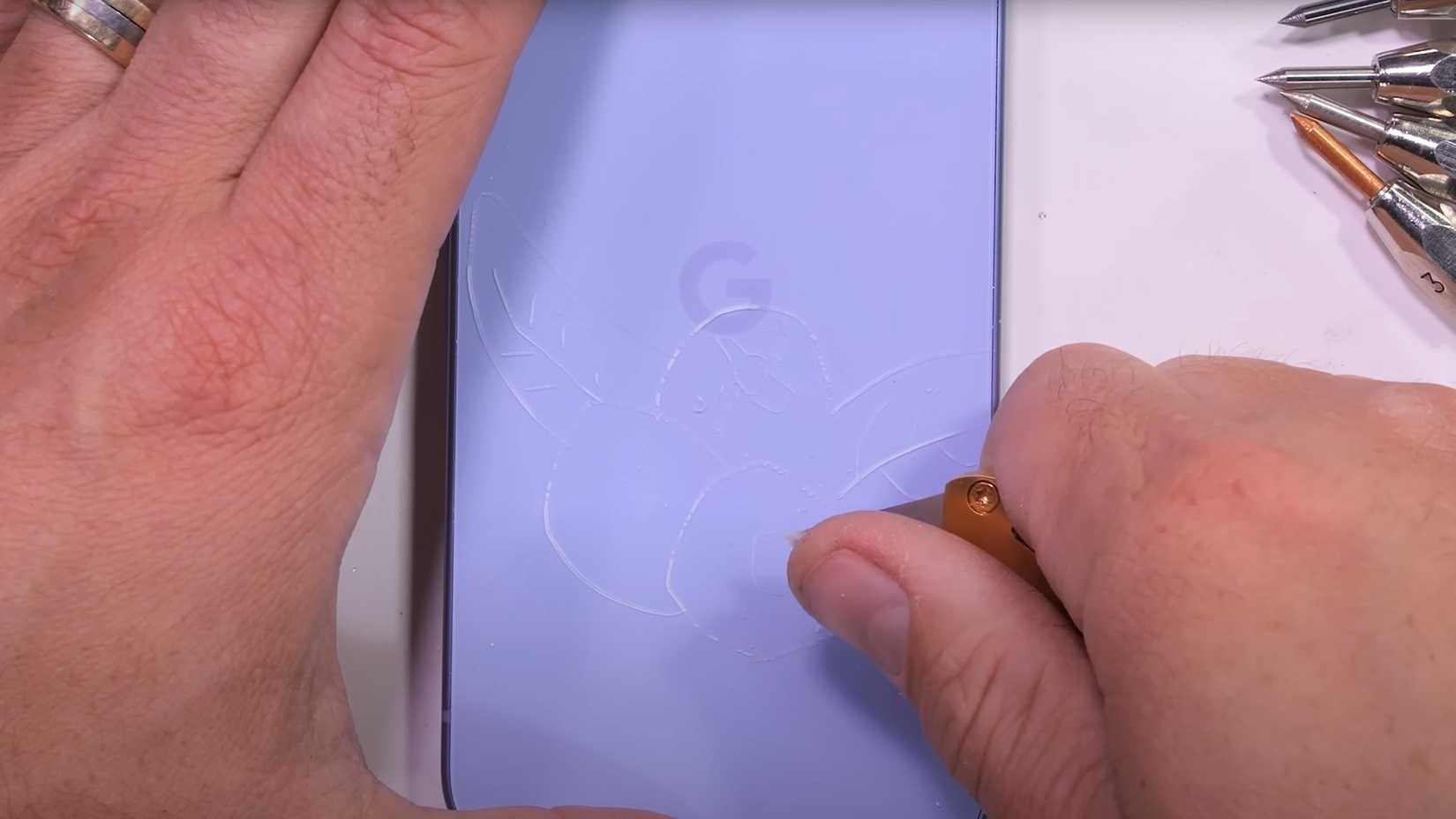 A screenshot of JerryRigEverything carving into the plastic back of the Pixel 9a with a razor knife.