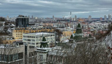 Panoramic winter view of Kyiv historic Podil district featuring distinctive green towers, colorful apartment buildings and dense urban landscape under dramatic cloudy sky with snow-covered rooftops.