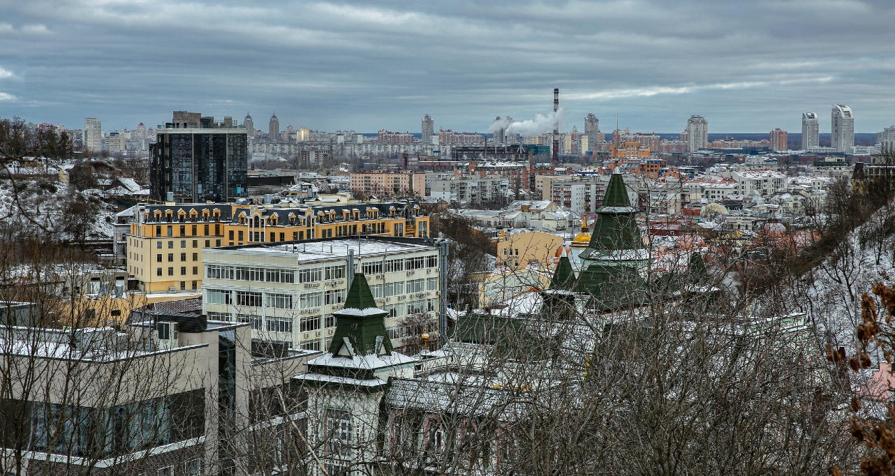Panoramic winter view of Kyiv historic Podil district featuring distinctive green towers, colorful apartment buildings and dense urban landscape under dramatic cloudy sky with snow-covered rooftops.