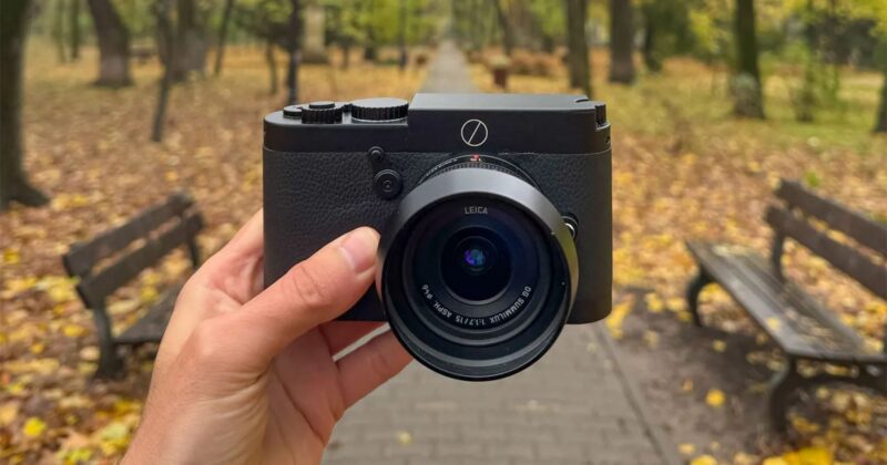 A hand holds a black Leica camera in the center of a park pathway lined with benches and autumn trees, with yellow leaves scattered on the ground.