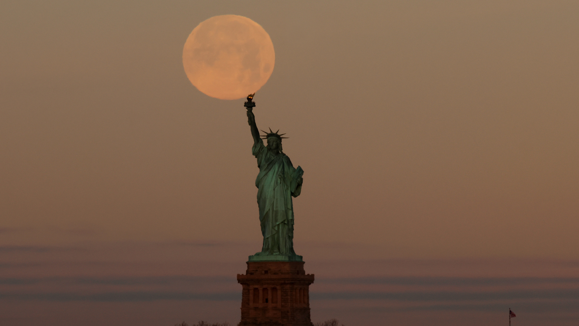 A yellow-orange full moon is pictured appearing to touch the torch of the Statue of Liberty in New York in a hazy evening sky.