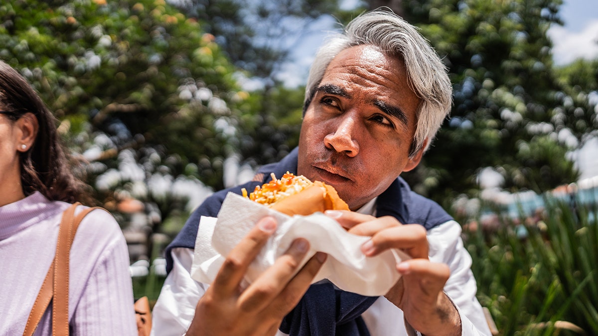 A man eats a loaded hot dog outside.