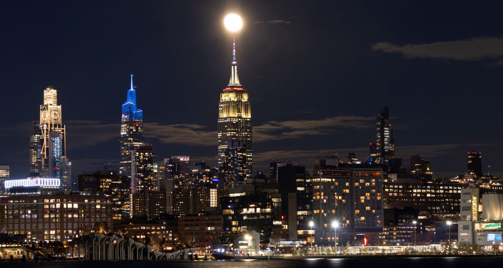A lit up cityscape at night, with various tall towers looking over smaller industrial buildings, with a waterfront in front of the city.