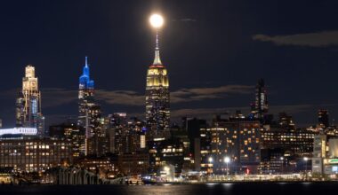 A lit up cityscape at night, with various tall towers looking over smaller industrial buildings, with a waterfront in front of the city.