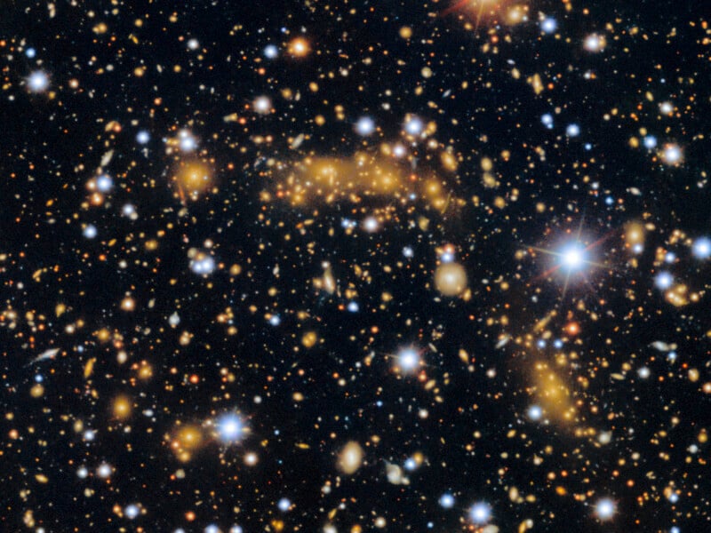 A dense field of stars and galaxies is scattered across the dark expanse of space. Some stars appear as bright points of light, while distant galaxies form faint, hazy shapes in shades of blue, orange, and yellow.
