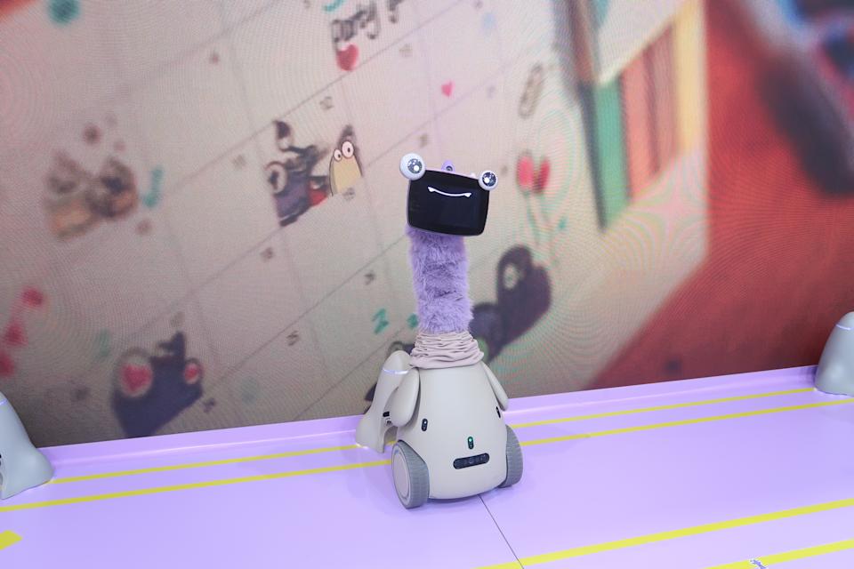 An OlloBot robot with a long purple neck stands in front of a display showing a home calendar covered in stickers