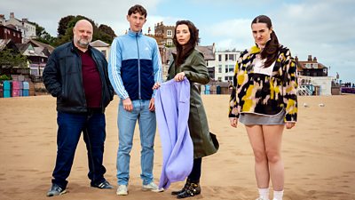Group image. Four people in differing attire stand on a sandy beach. A man on the left wears a leather jacket, jeans and trainers, while a younger man beside him wears a tracksuit jacket and jeans. The woman next to him wears a long coat and holds a purple pair of jogging bottoms. Stood slightly apart from the group is a young woman wearing a fleece and a long t-shirt. 