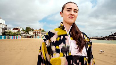 A young woman wearing a colourful fleece stands on a sandy beach. Colourful beach huts and a seaside town are visible in the background. 