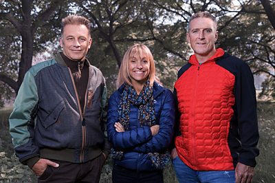 Two men (Chris Packham and Iolo Williams) stand in a forest with a woman (Michaela Strachan) in the middle - they are all wearing wintery clothing.