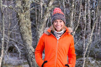 A woman in a wooly hat and orange coat (Michaela Strachan) smiles in a wintery forest 