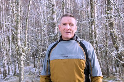A man in outdoorsy clothing (Iolo Williams) poses in a wintery forest 