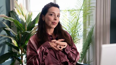 A woman in a maroon blouse sits behind a desk with her hands clasped together