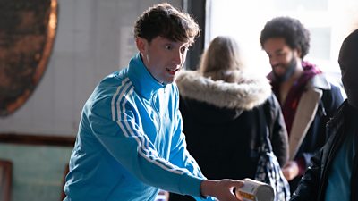A man in a blue tracksuit stands holding a can out to someone who's partially out of shot