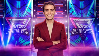 A man in a red suit smiles to camera as he stands with his arms folded, set against the backdrop of the Gladiators arena
