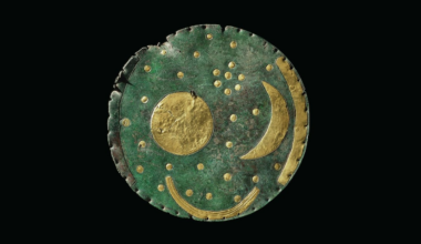 a green-patinaed bronze disk with gold accents representing a crescent moon, sun, and boat