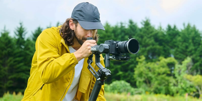 A person wearing a yellow jacket and black cap operates a professional camera on a tripod outdoors, with green trees and a cloudy sky in the background.