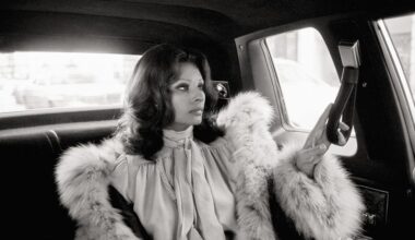 Sophia Loren is seen in a profile view sitting in the back of a car, wearing a voluminous fur-trimmed coat and a pussy-bow blouse.