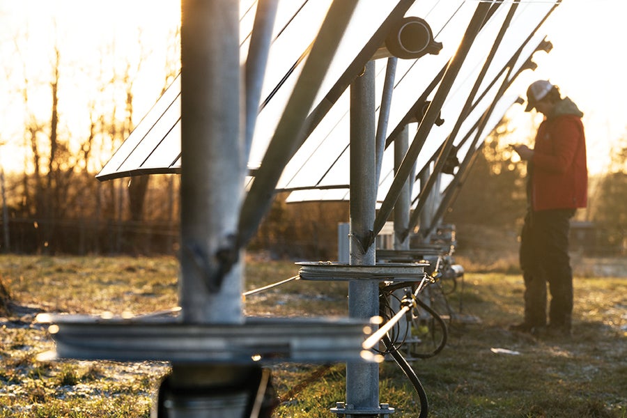 Close-up view down a line of vertical solar panel mounting posts. Under the panels, the posts pass through disks. A belt connects disks along the full series. A person in the background appears to be monitoring a motor connected to the belt.