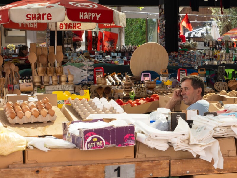 A man sits behind a market stall selling eggs, wooden kitchenware, and other goods, while talking on his phone. The stall is shaded by large red umbrellas that say "SHARE" and "HAPPY" in white letters.