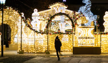 A person in winter clothing walks past festive holiday decorations made of bright yellow and white lights, featuring arches and fountains, on a city street at night. A man looks at her from behind.