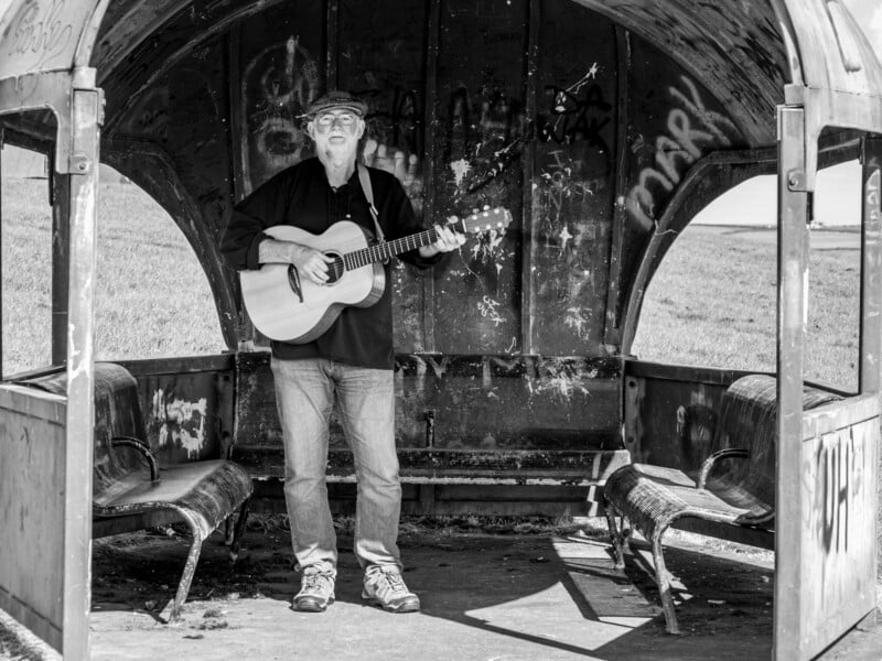 A man playing an acoustic guitar stands inside a weathered, graffiti-covered bus shelter with peeling paint and broken seats, surrounded by open grassy fields. The image is in black and white.