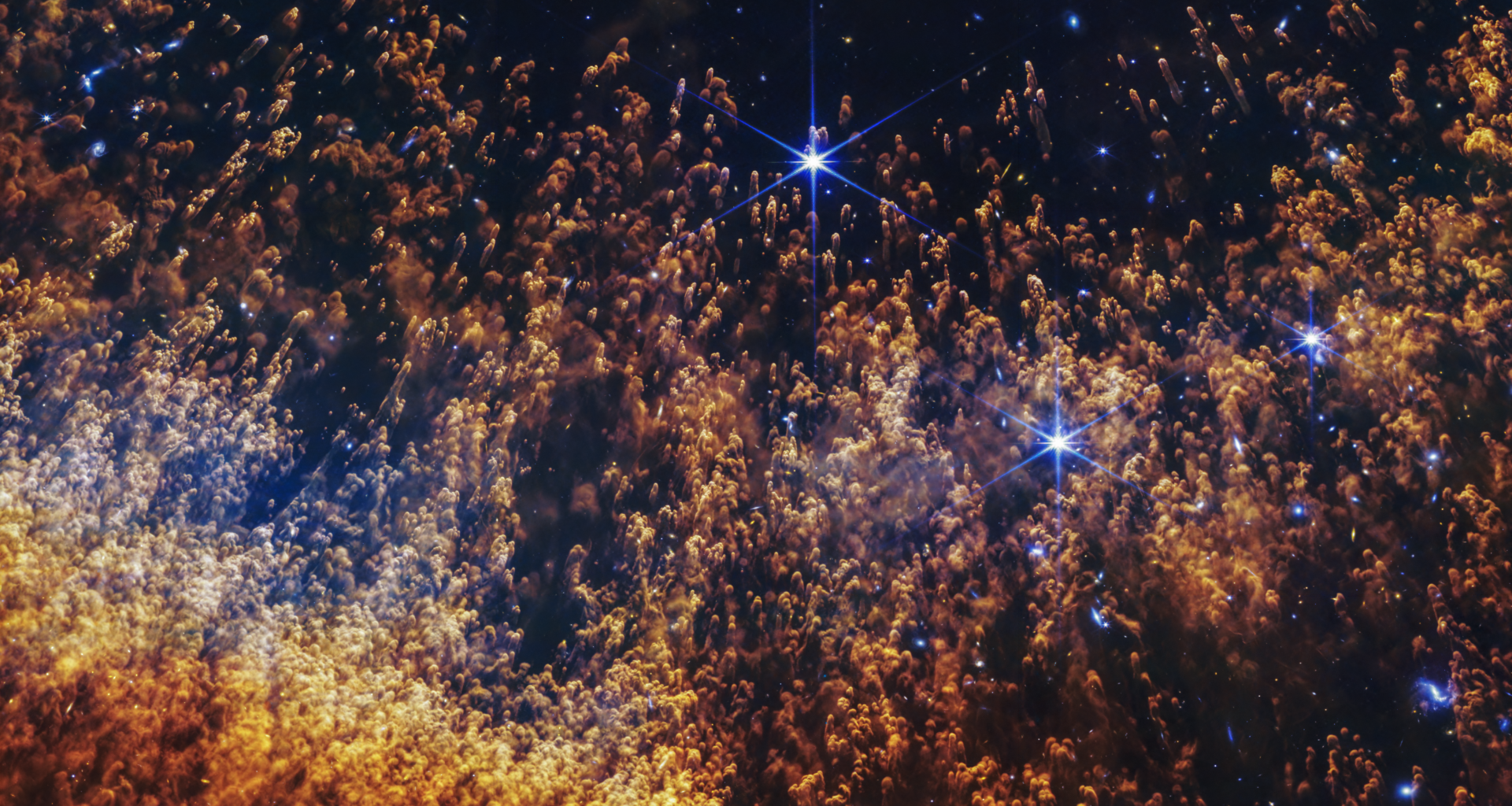A closeup of a small section of the Helix Nebula, an expanding shell of gas and dust. Thousands of orange and gold comet-like pillars stream upward from the bottom, like thin liquid blown up a sheet of glass. These pillars are around the circumference of the arced shell, which forms a partial orange semi-circle at the bottom. The pillars are more numerous and denser at the bottom, and darker red. They fade to orange and then yellow in the arc. In the top two-thirds, they are thinner and more golden, and it’s easier to see the black background of space. Several bright blue stars, some with diffraction spikes, are scattered throughout. A few larger stars are on the right side.