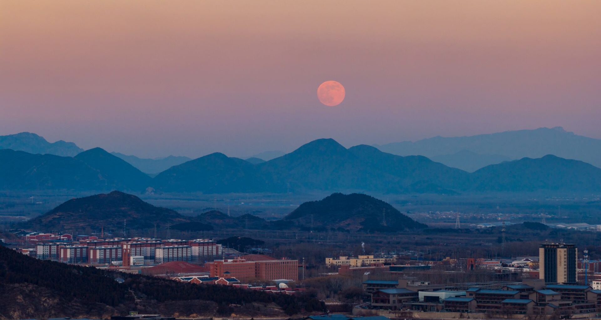 A full moon is pictured rising in the evening sky over a serene, misty mountain range bordering a city.
