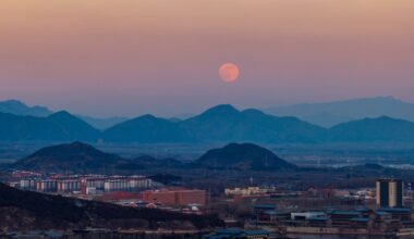 A full moon is pictured rising in the evening sky over a serene, misty mountain range bordering a city.