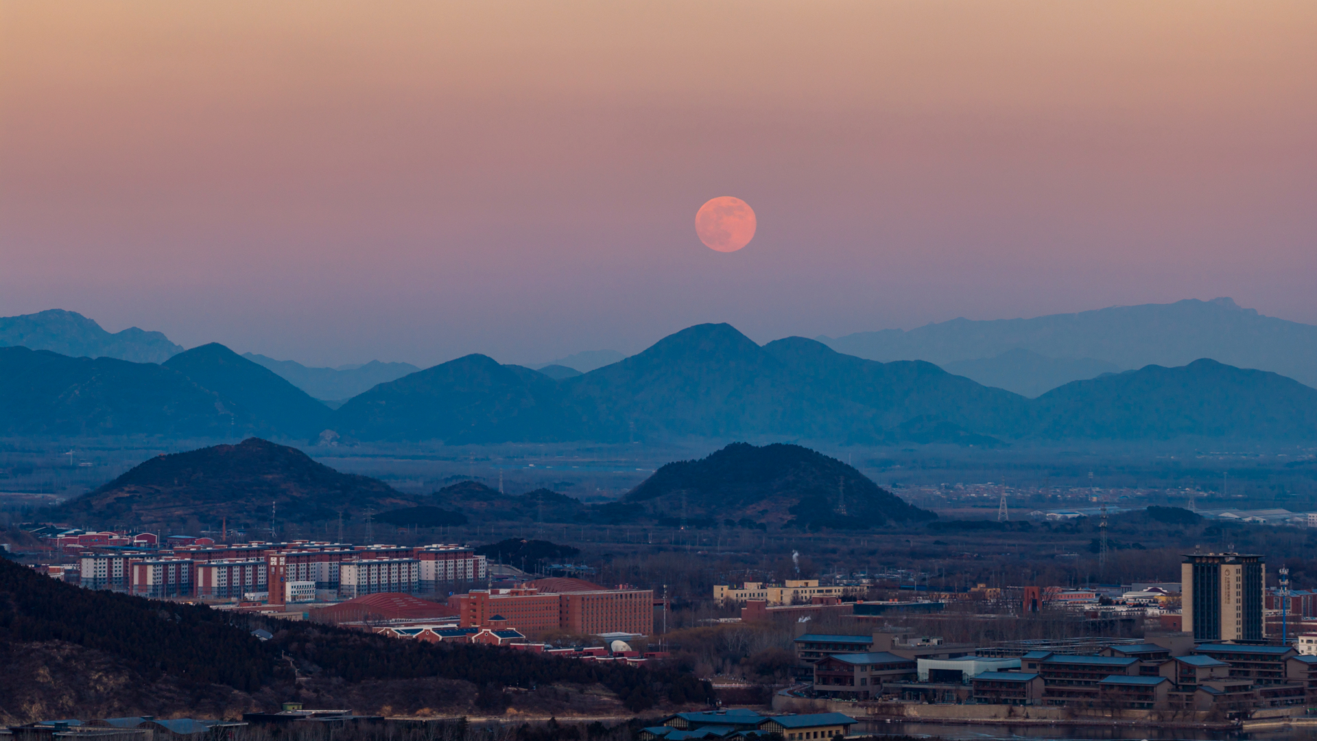 A full moon is pictured rising in the evening sky over a serene, misty mountain range bordering a city.
