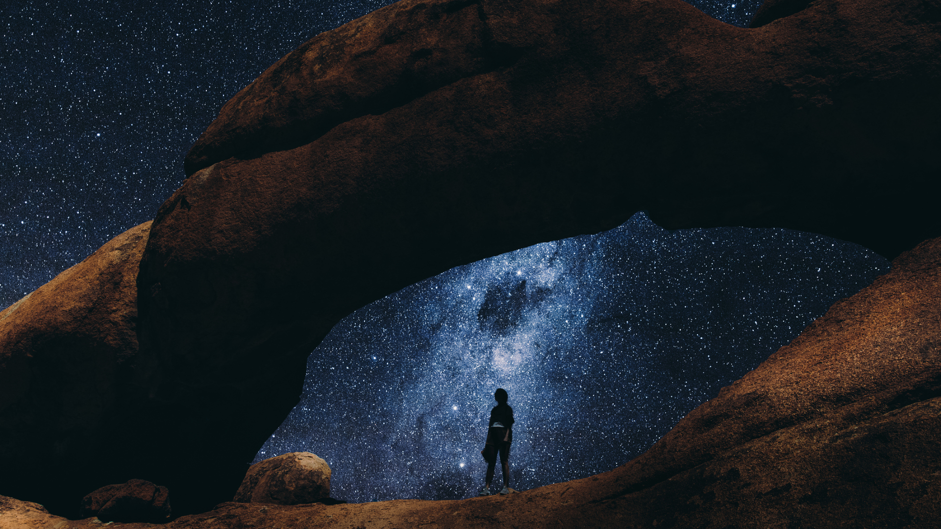 A silhouette of a person stands in between two large rocks with a dark blue starry night sky above them