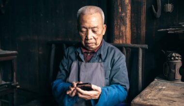An elderly man in a workshop setting, wearing a blue shirt and gray apron, sits focused on his smartphone against a backdrop of traditional wooden architecture and tools.