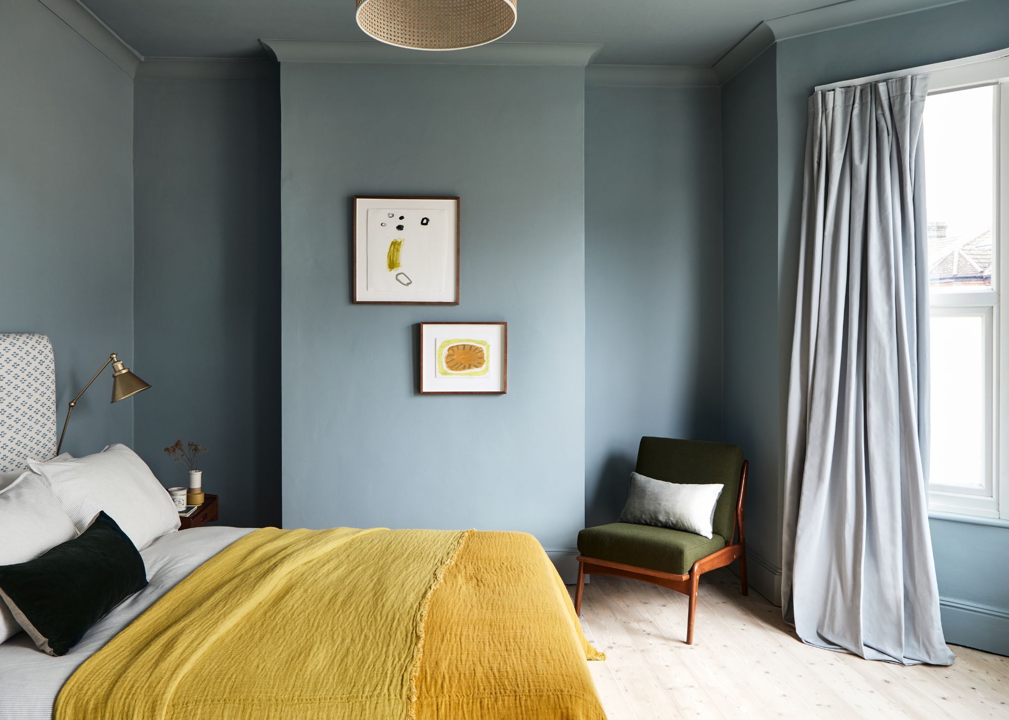 A bedroom with light blue walls, a bed with a yellow bedcover, and a bay window with pale blue curtains.
