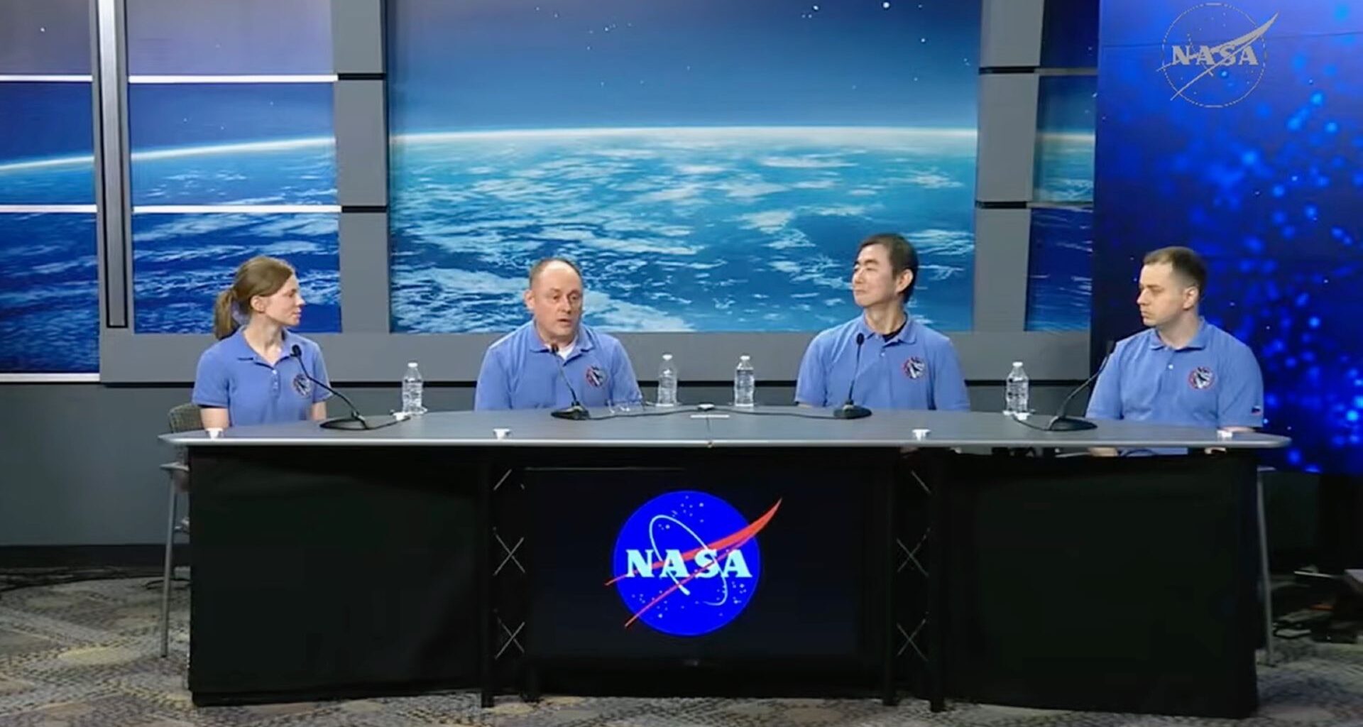 Four individuals wearing pale blue shirts sit behind a long table with a black cloth over it and the blue NASA circle logo with blue screens behind them.