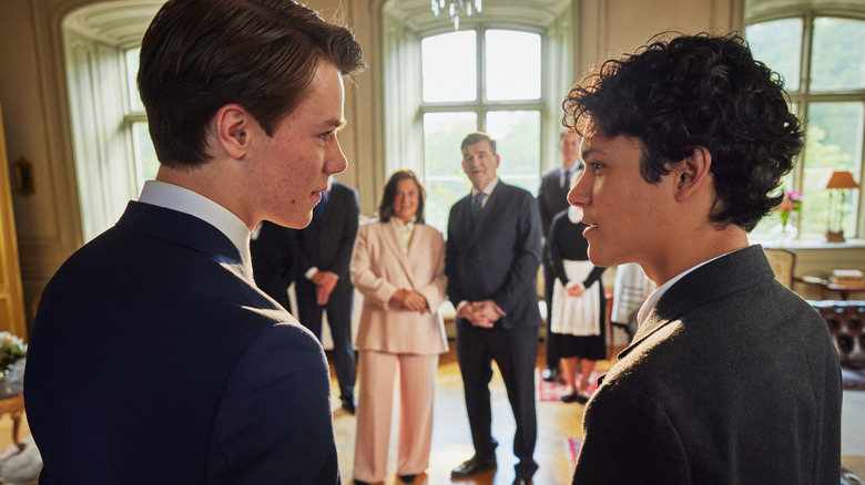 Edvin Ryding as Willhelm in suit standing next to and looking at Omar Rudberg as Simon with Pernilla August as Kristina and Magnus Roosmann as Ludvig in between them in the background in Young Royals