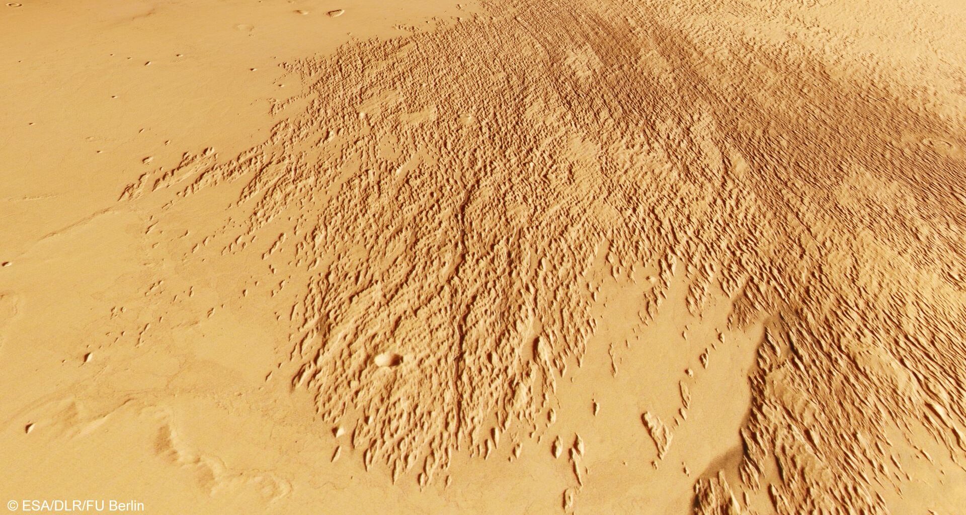 A section of Mars&rsquo;s surface showing a broad area covered with dense, elongated ridges and grooves that fan out diagonally across the image. The terrain is rough and textured in this region, contrasting with the smoother, lighter-coloured surrounding plains. The overall colour is a mix of tan and reddish-brown tones.