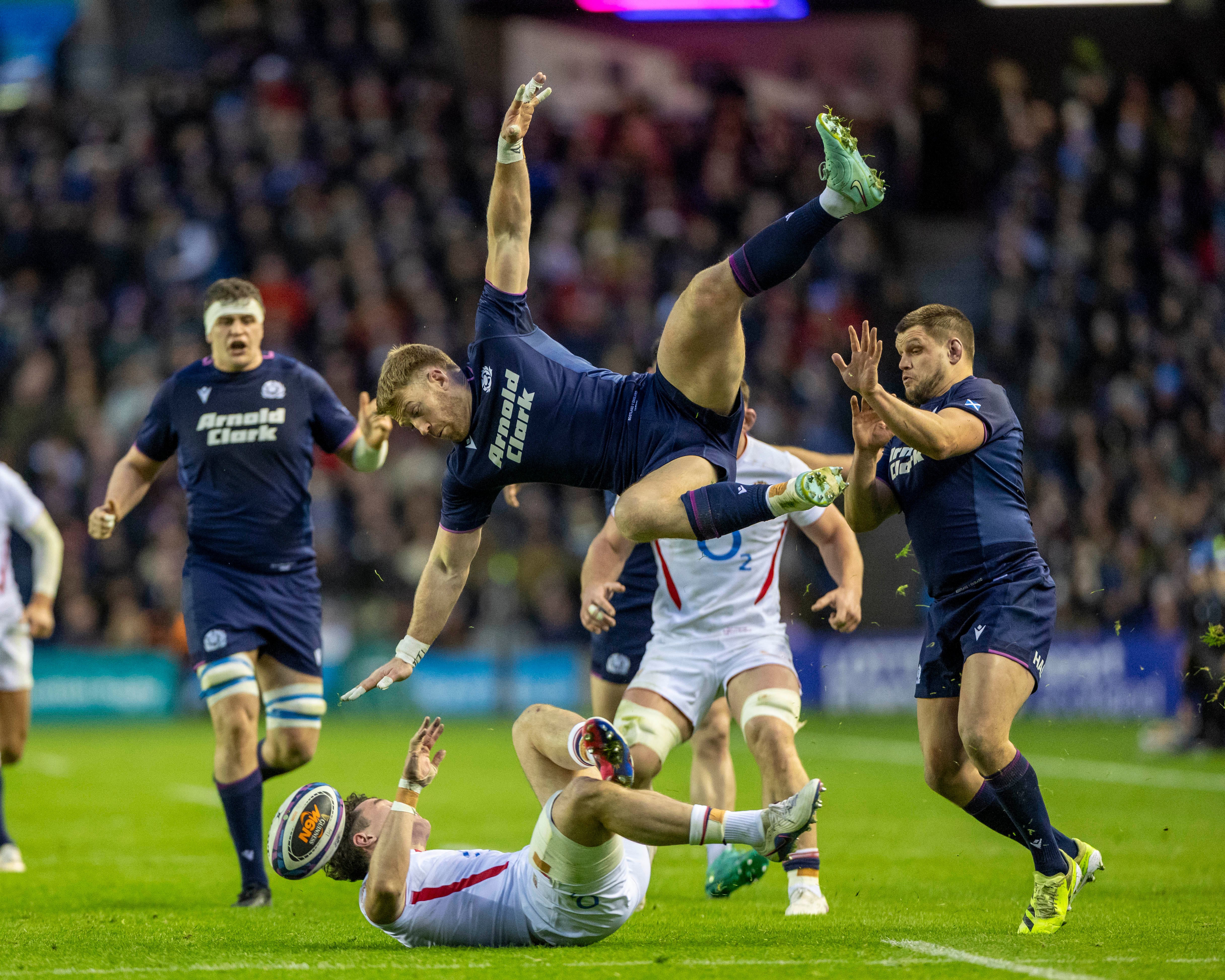 Rugby player Henry Arundell of England in a white jersey on the ground with a rugby ball, with Scotland player Kyle Steyn in a navy jersey mid-air.