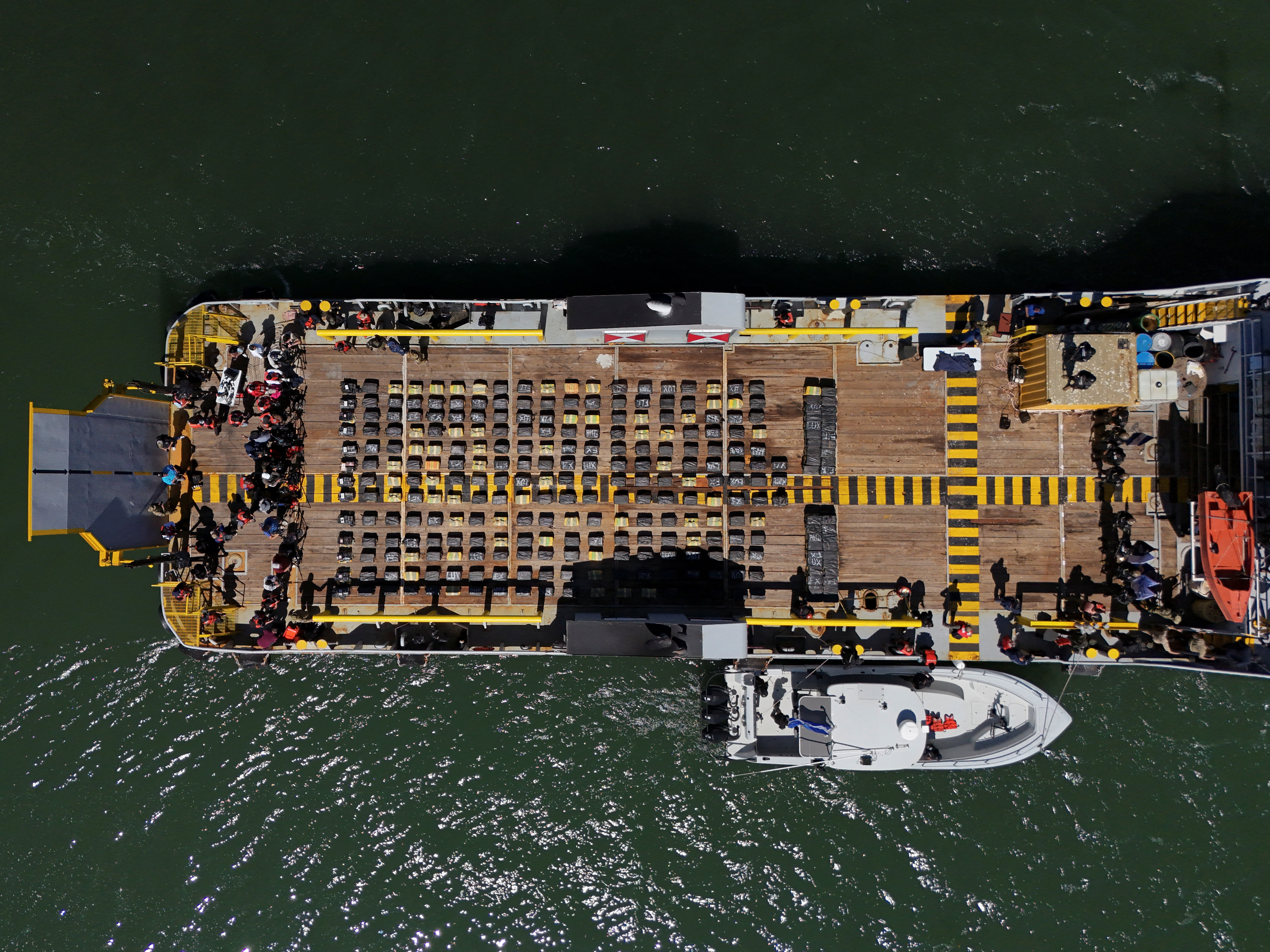 Drone view of security forces guarding large packages of seized cocaine on a vessel.