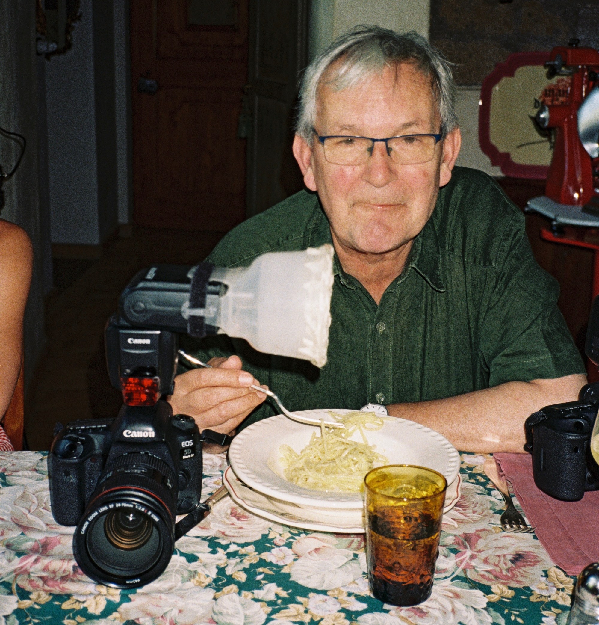A man in a green shirt and glasses eating pasta with a camera and flash next to him on a floral tablecloth.