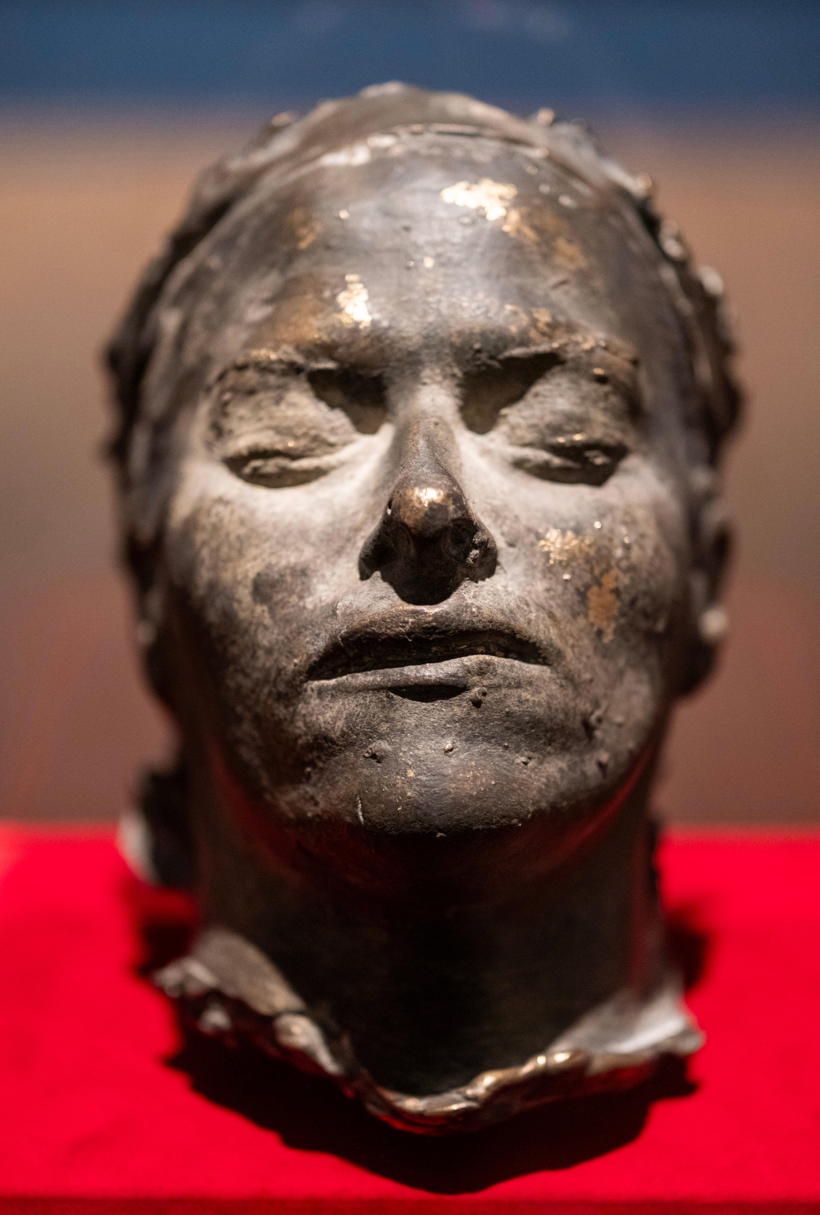 A death mask with closed eyes and mouth, with faint golden flecks on its bronze-like surface.