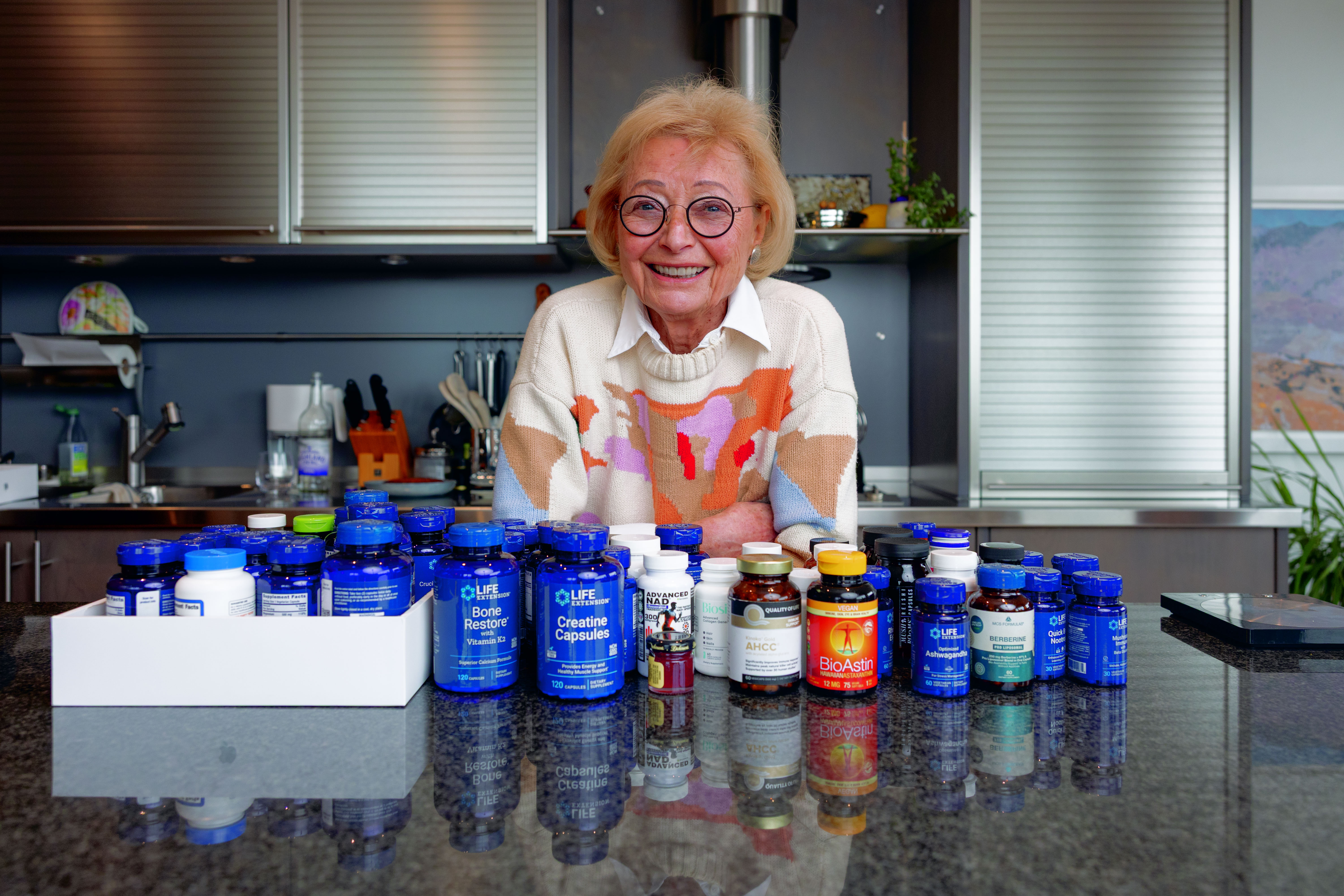 Helga Sands, an 88-year-old woman, smiles from behind a table covered with various supplement bottles in her kitchen.
