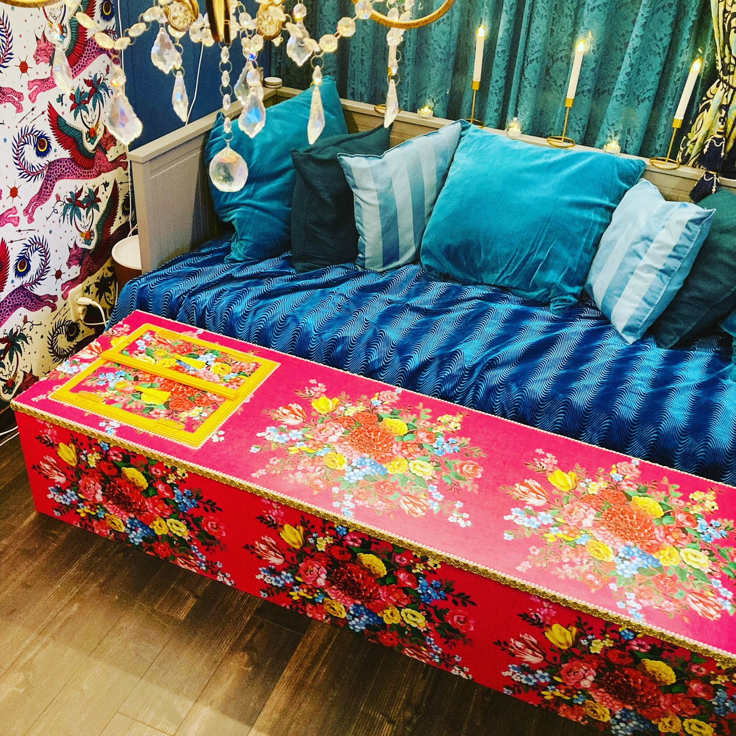 A brightly colored coffin with a floral pattern, in a room with a blue sofa and patterned wallpaper.
