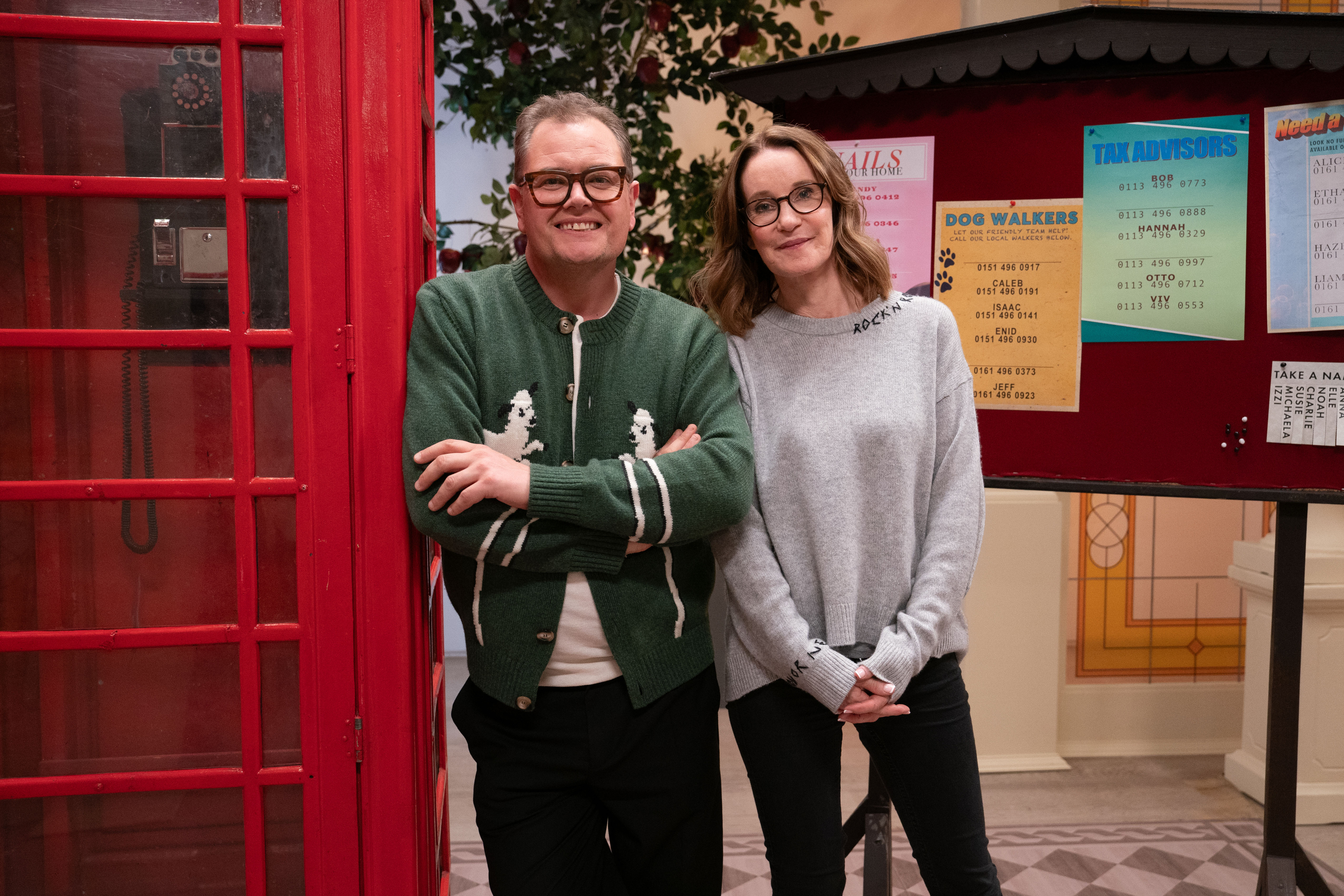 Alan Carr and Susie Dent stand next to a red phone booth and a bulletin board.