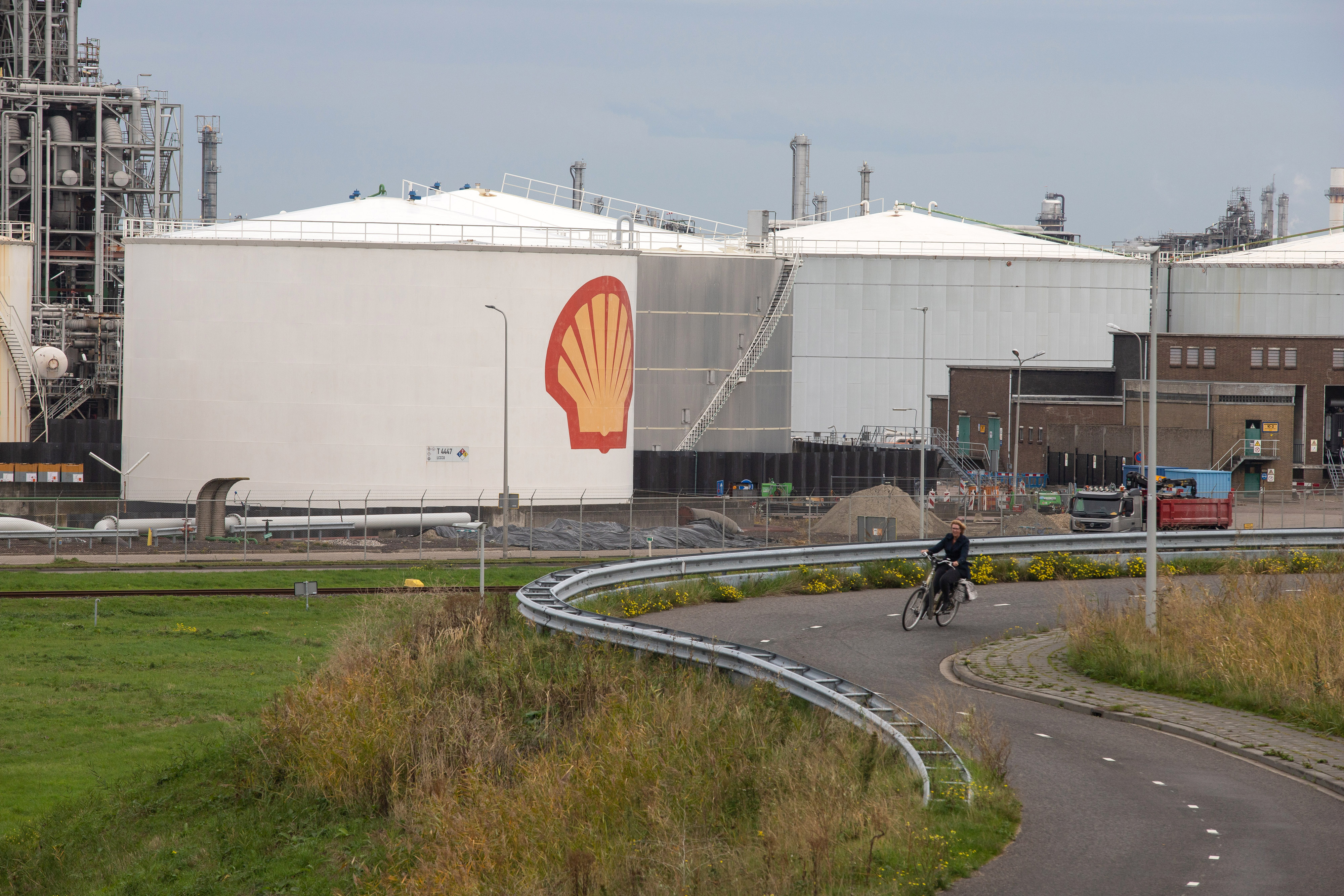 A cyclist on a bike path passes by oil storage silos and refinery infrastructure.