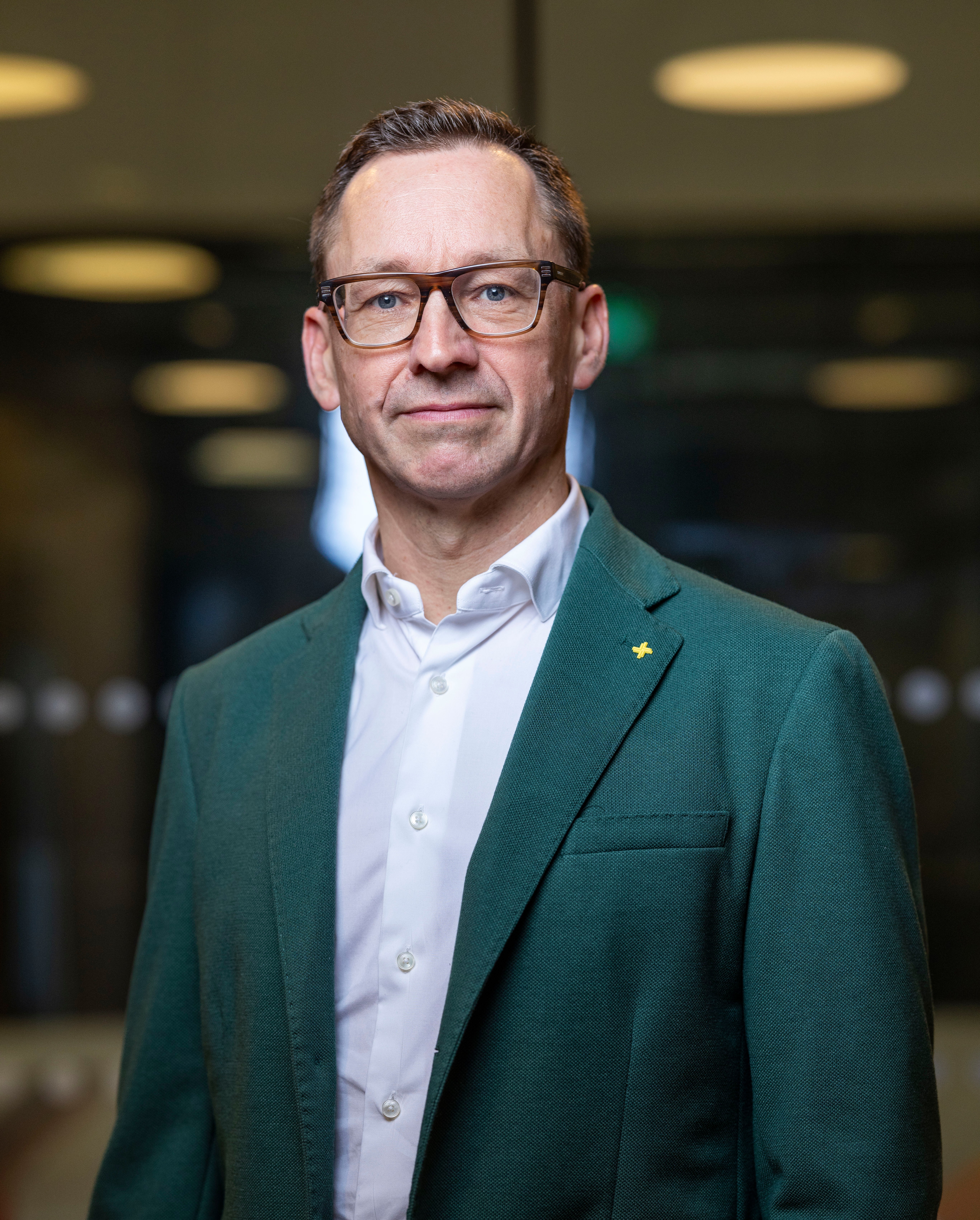Malcolm Craig, a man with short brown hair, wearing glasses and a green jacket with a white collared shirt, is looking directly at the camera.