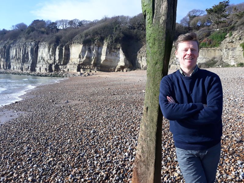 Andy Dinsdale standing on a pebble beach with cliffs in the background.