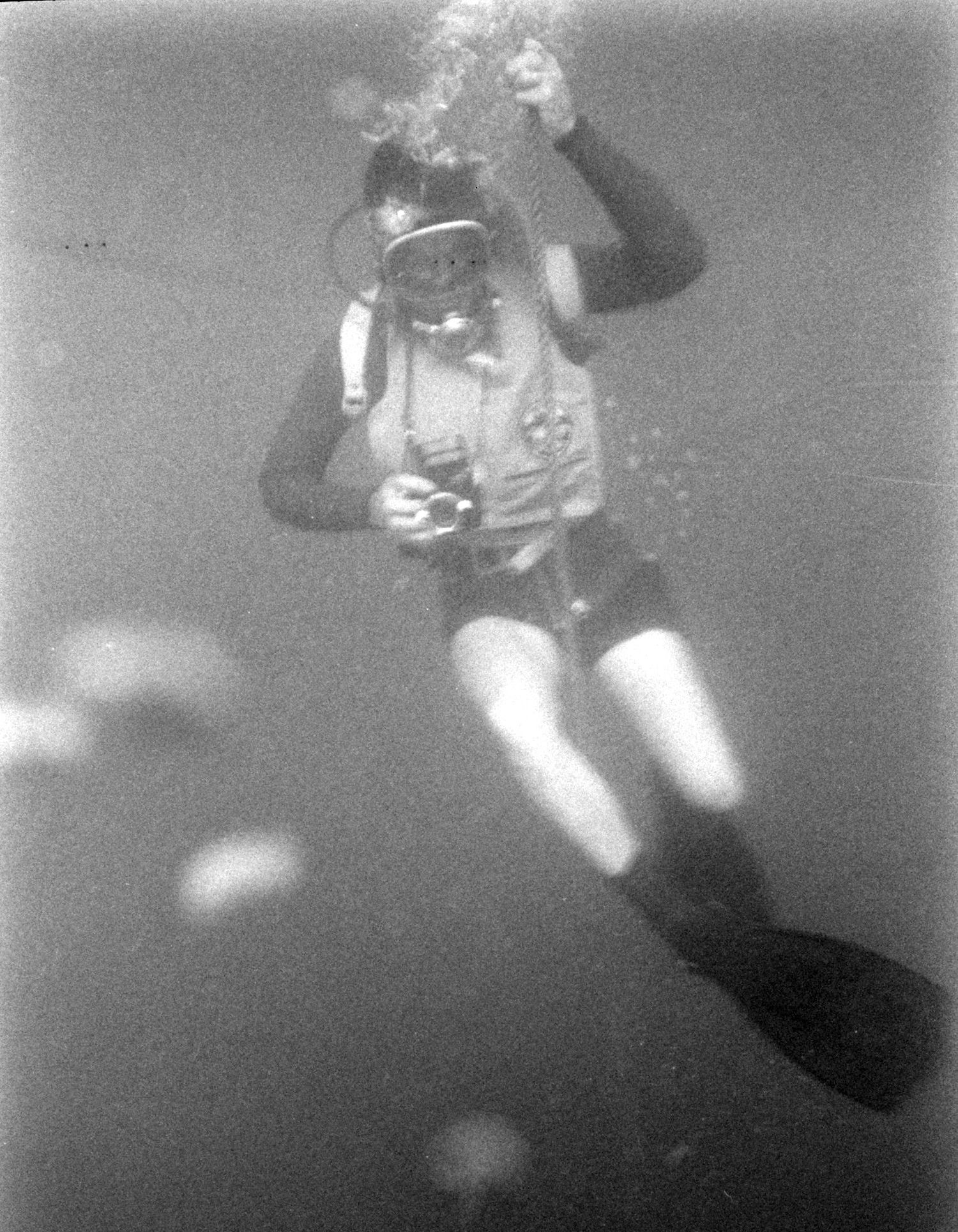 Underwater diver with a camera, wearing a mask, wetsuit, flippers, and a buoyancy compensator.